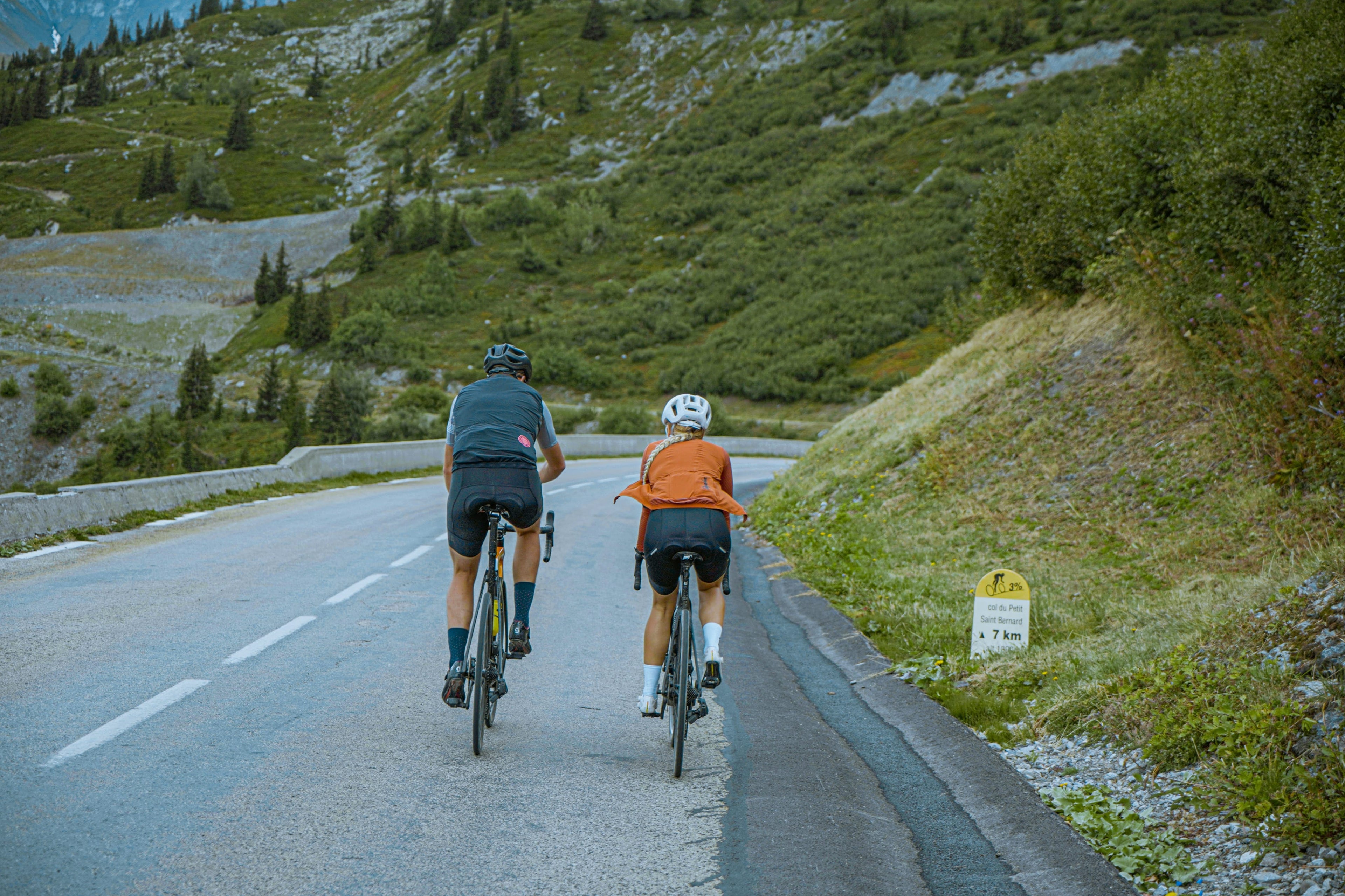 Two cyclists riding on a mountain road with greenery and a sign in the background.