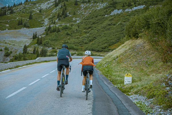 Two cyclists riding on a mountain road with greenery and a sign in the background.