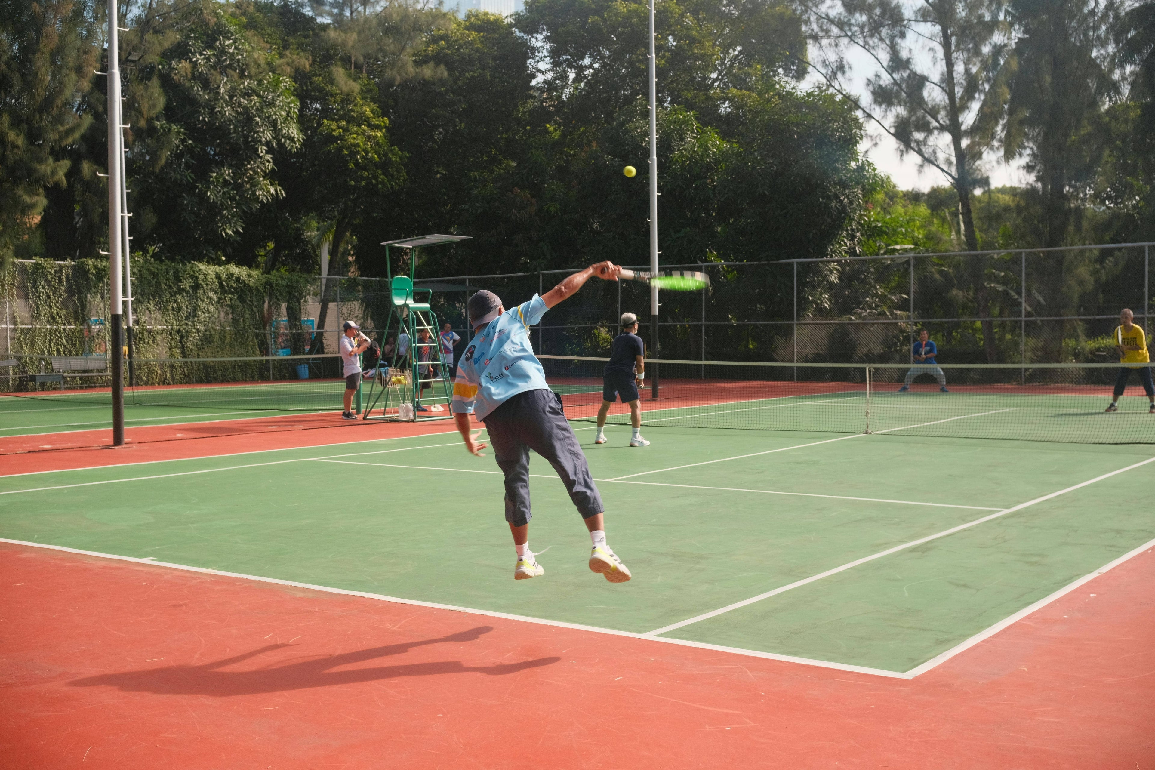 Person playing tennis on a red and green court with trees in the background