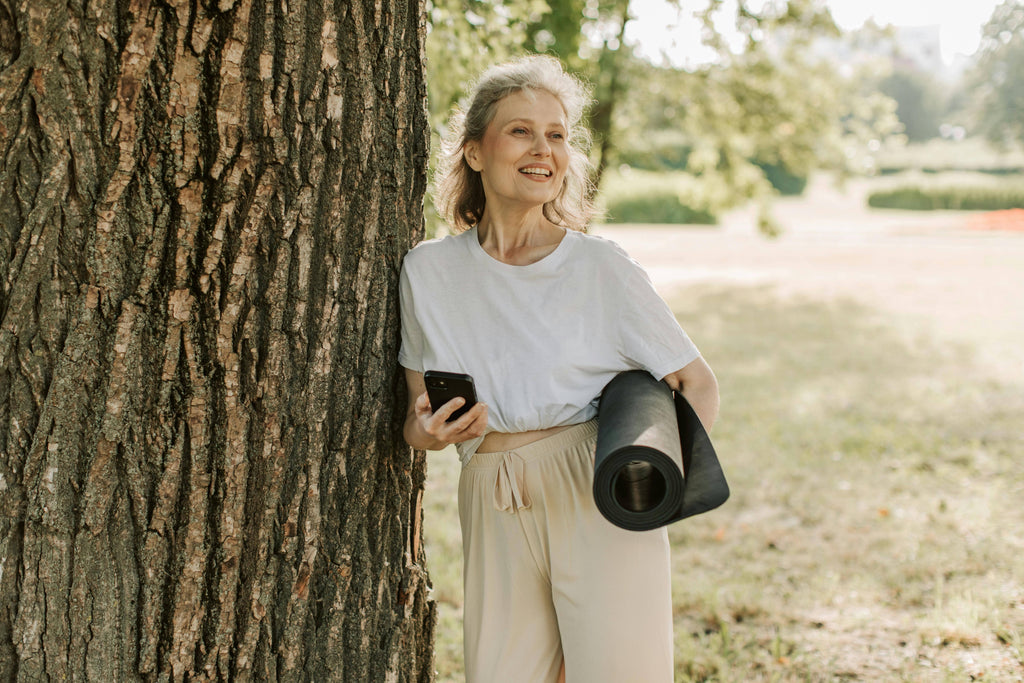 Woman holding a phone and yoga mat leaning against a tree in a park