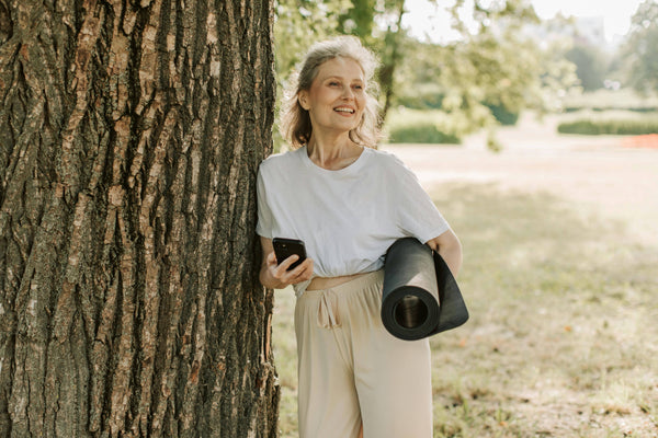 Woman holding a phone and yoga mat leaning against a tree in a park